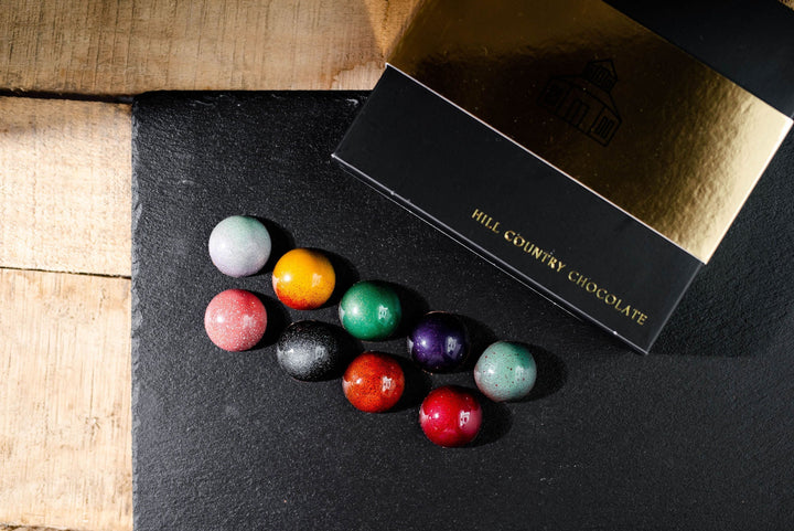 A black and gold Hill Country Chocolate box sits beside nine vibrant, glossy Signature Artisanal Bonbons—a handcrafted collection from Hill Country Chocolate—displayed on a dark slate surface.