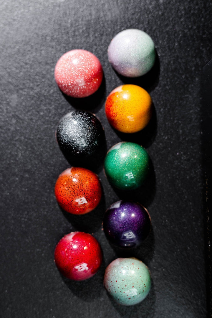 Nine glossy, colorful bonbons from Hill Country Chocolate’s 9 Piece Signature Artisanal Bonbons collection are arranged in three vertical columns on a dark background—a true delight for chocolate lovers.