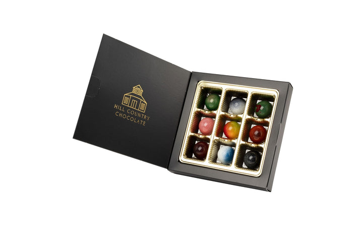 An open black box labeled "Hill Country Chocolate" displays a handmade collection of nine assorted, colorful Signature Artisanal Bonbons in a gold tray—perfect for chocolate lovers.