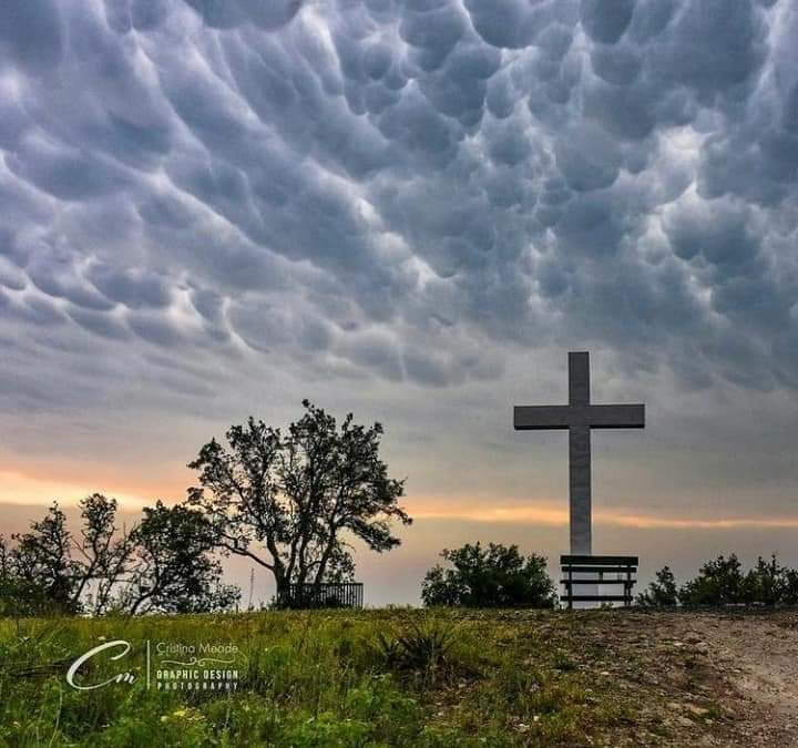 Cross Mountain by Cristina Mead, large white stone cross on grassy hill with dramatic cloudy sky at sunset