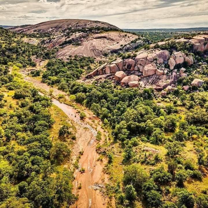 Aerial view of Enchanted Rock in Fredericksburg, Texas, featuring pink granite dome, large boulders, green trees, and a natural stream.