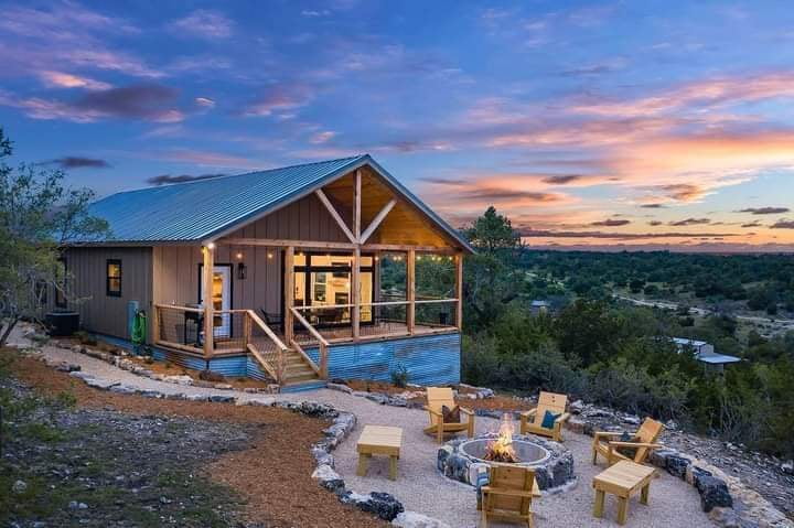 Rustic wood cabin with metal roof, outdoor fire pit, and wooden chairs at sunset near Fredericksburg