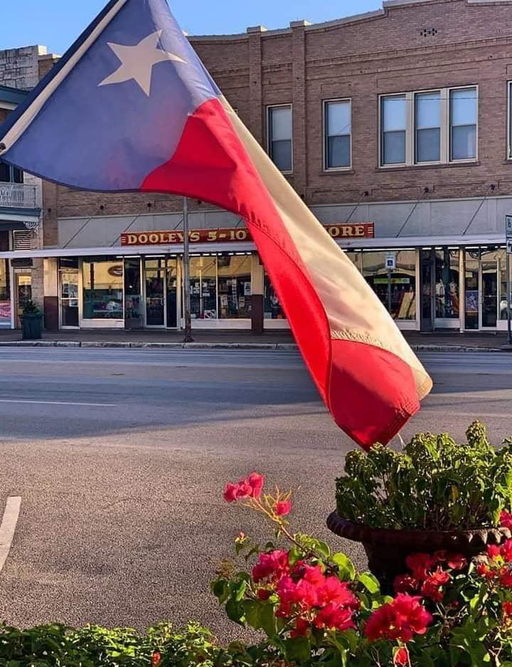 Texas flag in red, white, and blue fabric flying by blooming pink flowers, downtown Fredericksburg