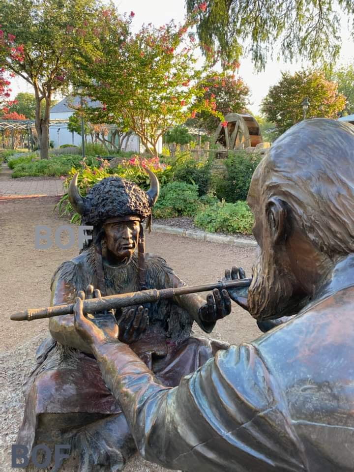 Bronze outdoor sculpture of two figures, one in a horned headdress, sharing a peace pipe