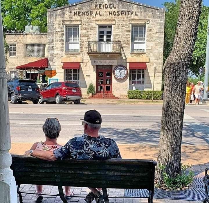 Couple sitting on a bench under a tree, facing historic Keidel Memorial Hospital stone building.