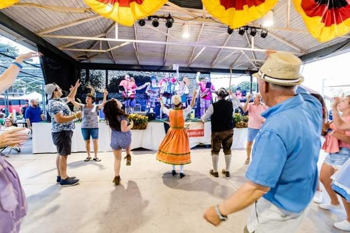Outdoor festival with people dancing in colorful outfits, live band on stage, festive decorations