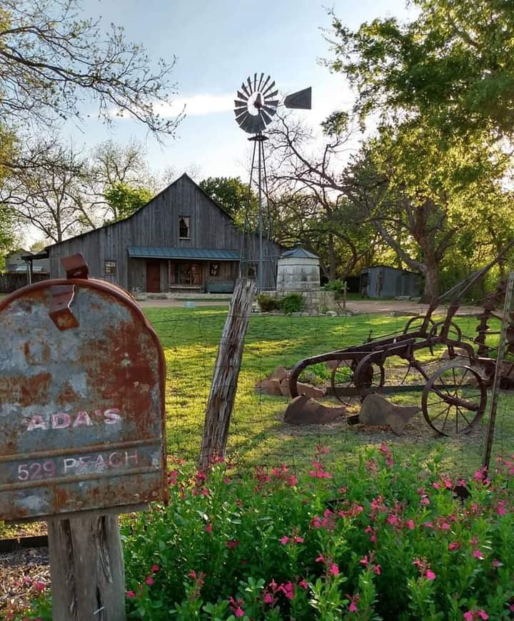Rustic metal mailbox, vintage farm plow, windmill, and barn in a grassy Texas homestead scene.