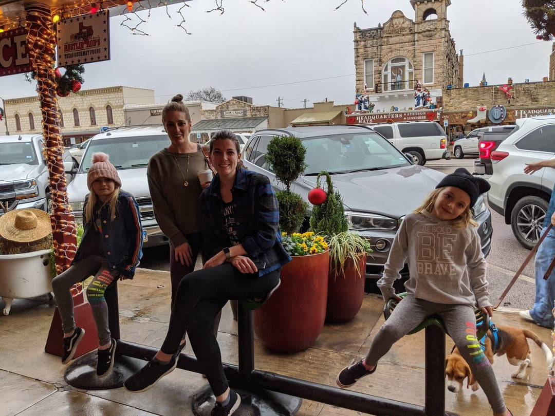Two girls in rainbow-striped pants and BE BOLD BE BRAVE sweatshirt, denim jacket, knit hats, posing with women and dog outside boutique, planter decor visible