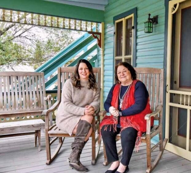 Two women sitting on wooden rocking chairs on a turquoise porch with decorative trim, one in a beige sweater dress and boots, the other in a navy top with red scarf.