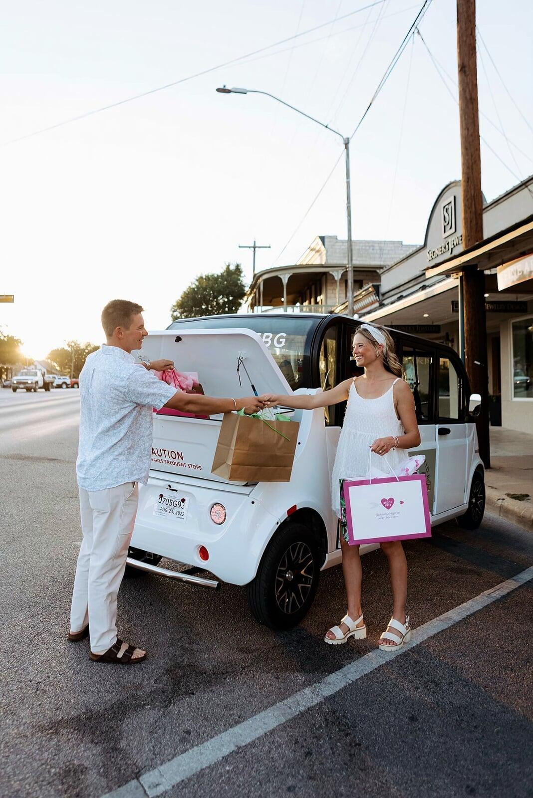 Two people exchanging shopping bags outside a white electric vehicle on a Fredericksburg street, featuring large brown and pink paper gift bags.
