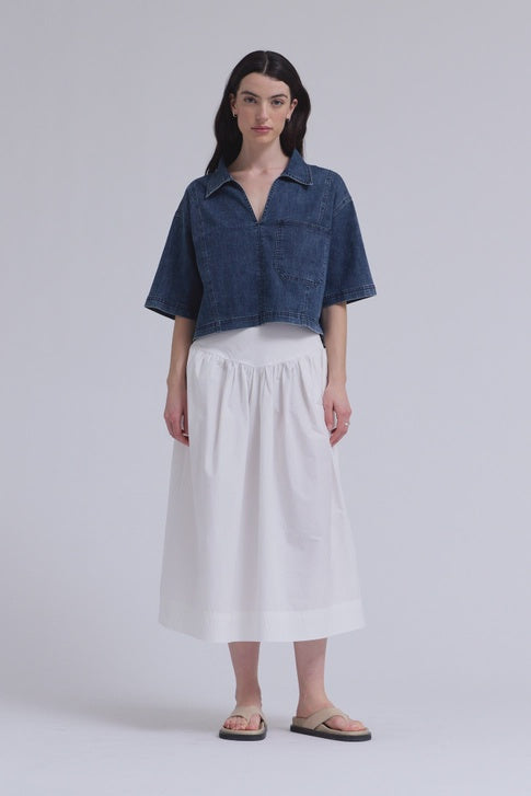A model with long dark hair and a neutral expression wears the Zertz DENIM CROP SHIRT, paired with a white mid-calf skirt and beige sandals against a plain background, effortlessly showcasing this versatile wardrobe essential.