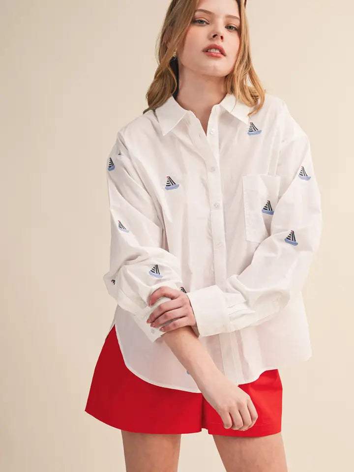 A woman wears the BOAT EMB TOP by Zertz—a loose white classic top with small blue triangle patterns—and bright red shorts, standing against a plain beige background.