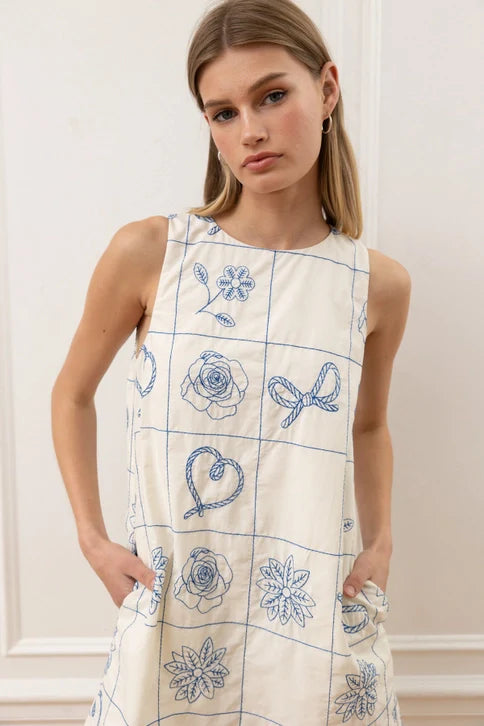 A woman with straight blonde hair models the Zertz FLORAL PATTERN SLVLESS MINI DRESS, a white sleeveless dress featuring blue floral embroidery and pockets, standing against a light-colored wall.