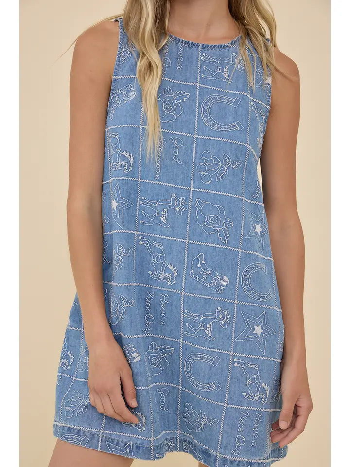 A person wears the Zertz EMBROIDERED DENIM WESTERN DRESS, featuring sleeveless blue denim with white stitched cartoon stars, horseshoes, and flowers for a playful cowgirl look against a beige background.