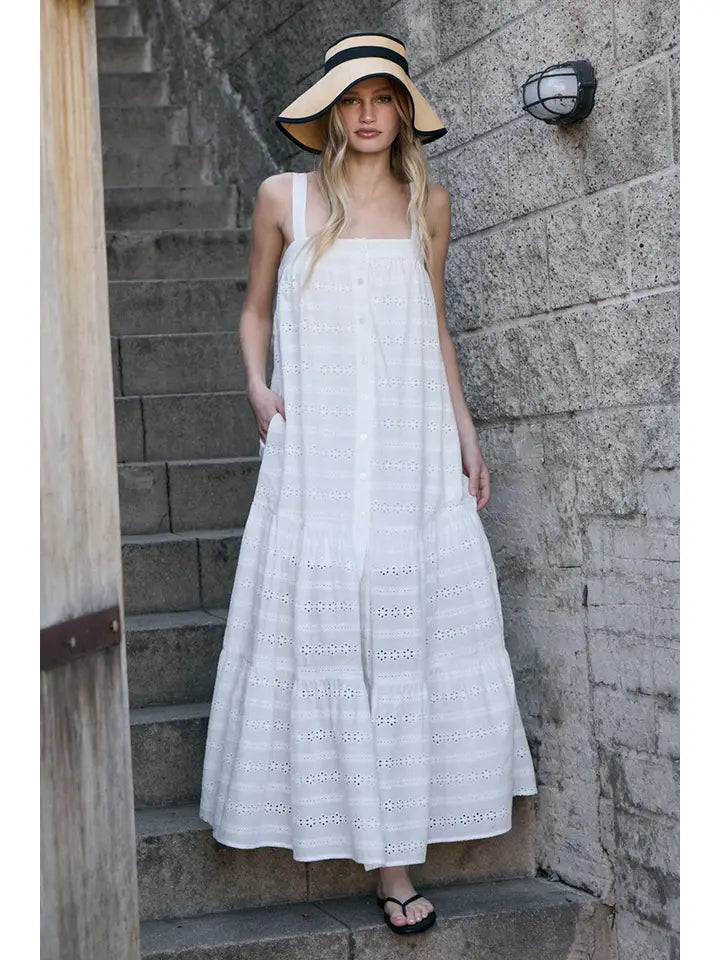 A woman with long blonde hair, wearing the Zertz EYELET BUTTON FRONT MAXI DRESS and a wide-brimmed straw hat, stands on outdoor concrete steps by a stone wall in sandals.