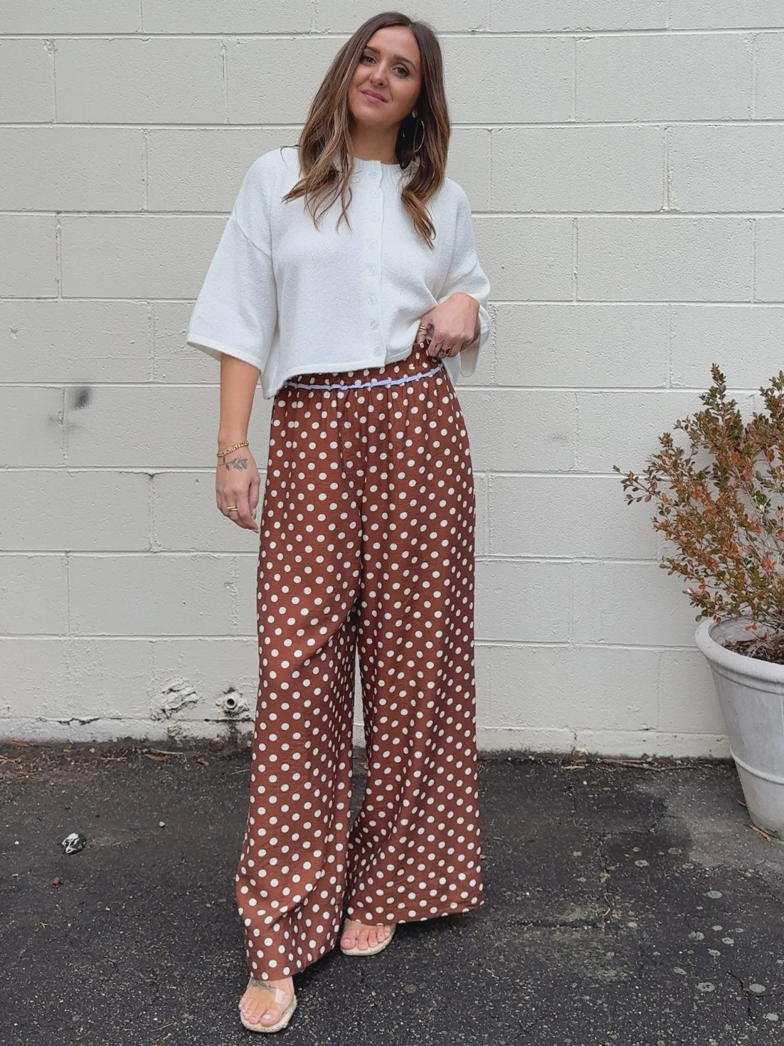 A woman smiles in front of a white brick wall, wearing a white buttoned top and Zertz POLKA DOT PANTS—high-waisted, wide-leg brown pants with playful polka dots. She stands with one hand on her hip beside a potted plant.