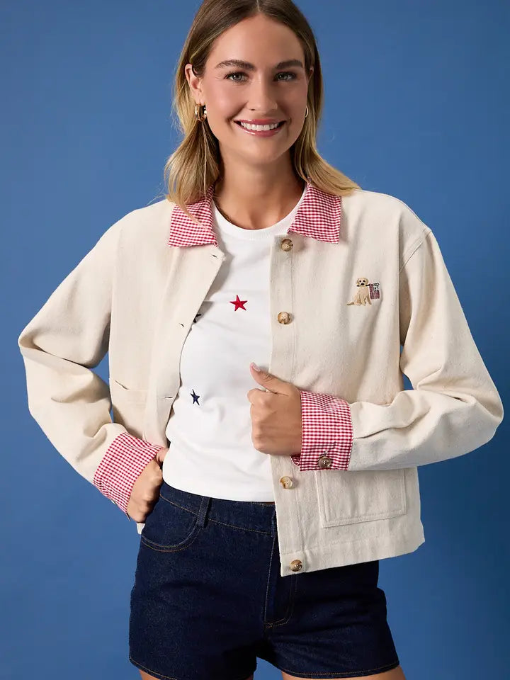 A woman stands against a blue background, smiling with hands on her hips. She wears the Zertz USA FLAG BARN JACKET featuring a red gingham collar and cuffs, paired with a star-print white t-shirt and dark shorts.
