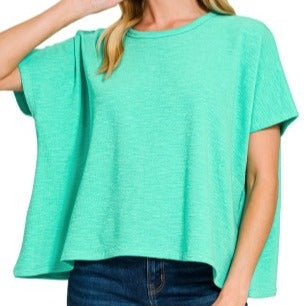 A woman wears the Loca on Main Cotton Club Tee—a loose, short-sleeved, mint green top with a relaxed fit and round neckline—paired with dark blue jeans.