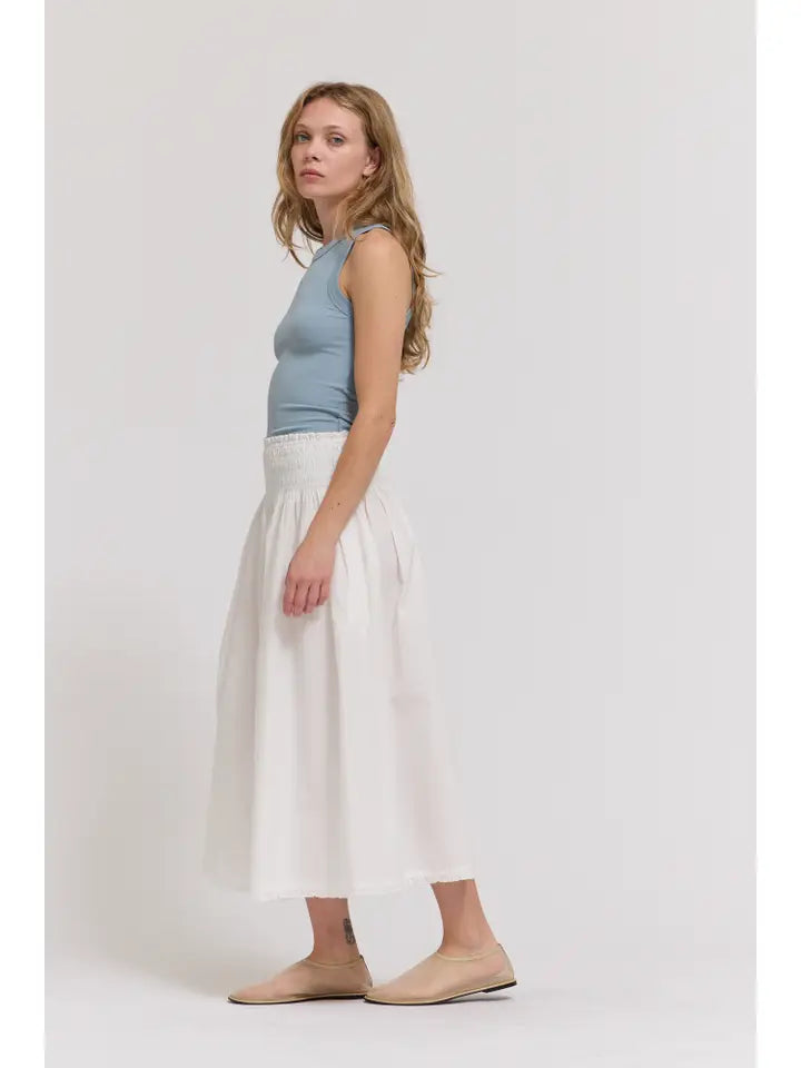 A woman stands sideways against a white background wearing a light blue sleeveless top, beige flats, and the Zertz SMOCKED WAIST SKIRT with eyelet trim.