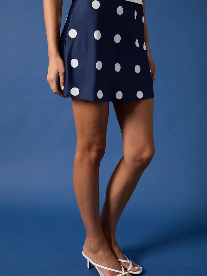 Only the lower body is visible as a person stands against a blue background, wearing the Zertz MINI SATIN SKIRT in navy blue with large white polka dots and white heeled sandals.