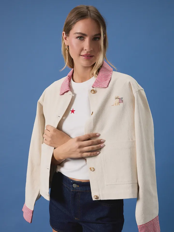 A woman poses against a blue backdrop wearing the Zertz USA FLAG BARN JACKET, a cream durable canvas jacket in a relaxed fit with red gingham details, over a white shirt with a red star and dark shorts, holding the jacket slightly open.