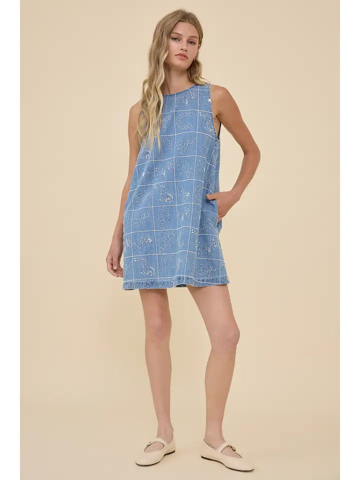 A woman models the Zertz EMBROIDERED DENIM WESTERN DRESS, featuring white checks and floral embroidery. She pairs it with light loafers, her long wavy blonde hair enhancing the cowgirl-inspired look against a beige backdrop.