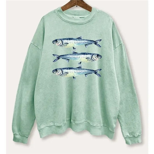The Three Sardines Sweatshirt by Loca on Main is a light green, oversized 100% cotton top with three realistic fish illustrated across the chest. Shown on a wooden hanger against a neutral background.