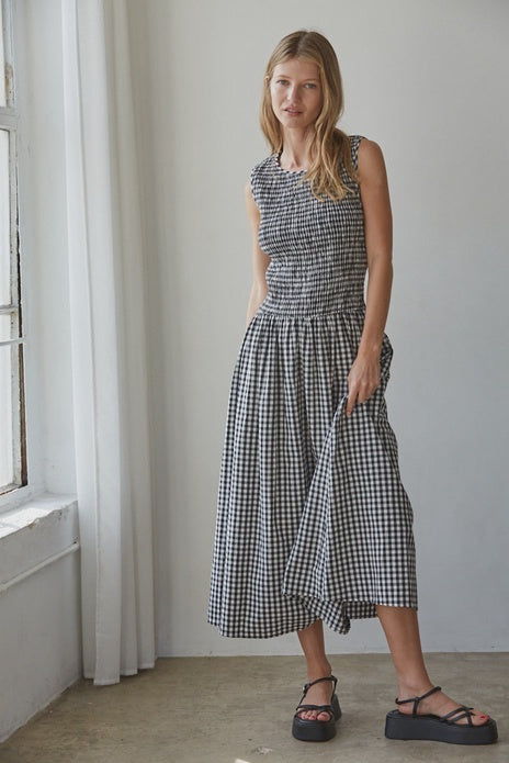 A woman stands indoors by a window wearing the Zertz SAYLER GINGHAM SMOCKED DRESS, a sleeveless cotton midi with a gathered waist, paired with black sandals. She holds her skirt and looks at the camera; white curtains hang nearby.