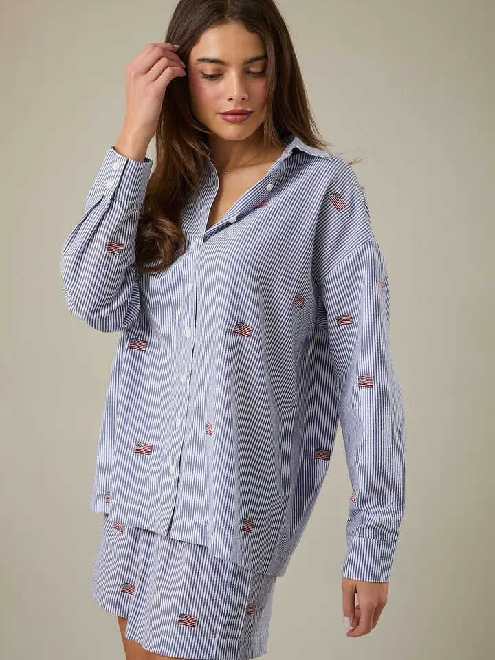 A woman with long brown hair models the Zertz STRIPE FLAG BUTTON DOWN TOP, featuring blue and white stripes with small flag patterns, paired with a matching skirt against a plain background—a true wardrobe essential.