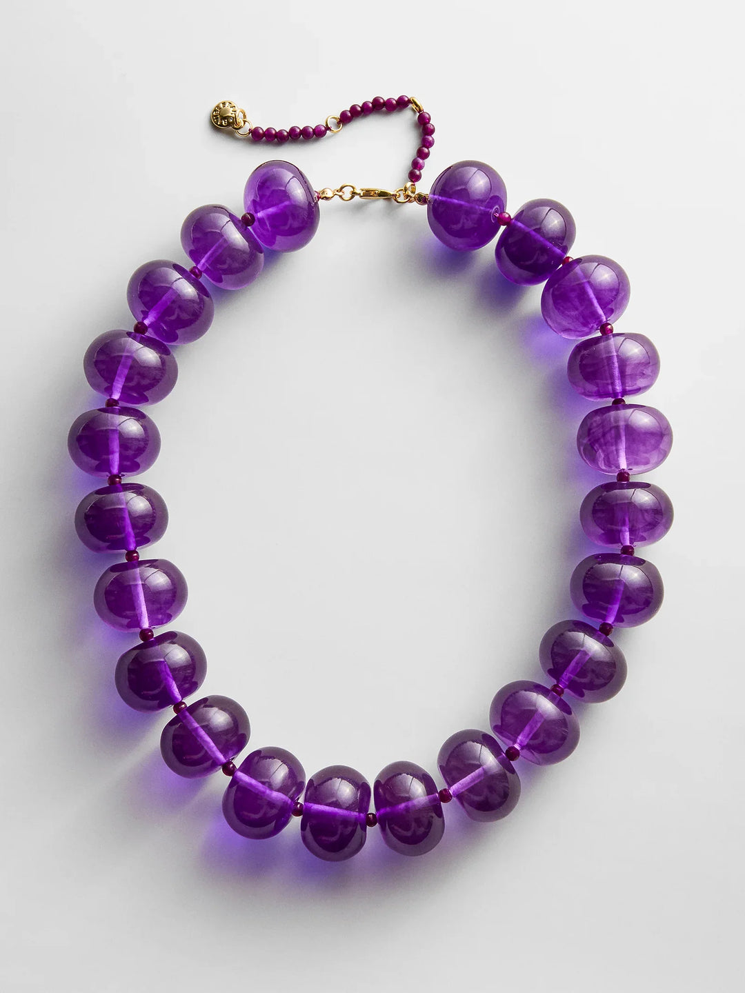 The JOAN SEMI PRECIOUS NECKLACE by Zertz features large, round, translucent purple beads with smaller purple beads and a gold clasp, beautifully arranged in a circle on a white background.