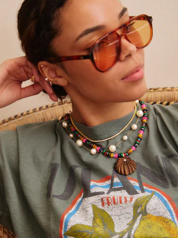 A woman wearing large orange sunglasses, a green fruit-print graphic t-shirt, and the Zertz SUN DRIFT SEMI-PRECIOUS PNDT NECK necklace with statement beads and pearls, sits on a wicker chair.