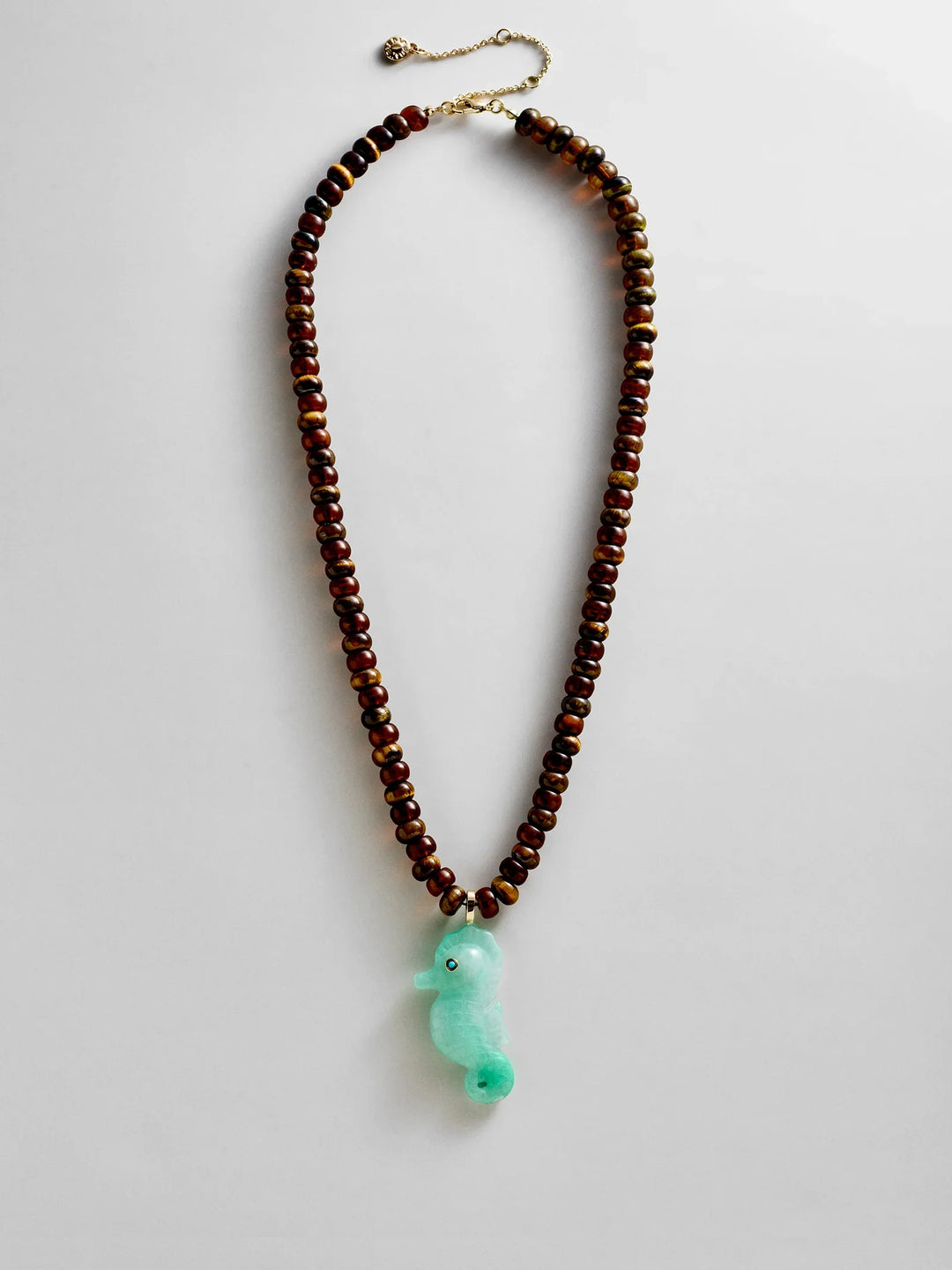 The Zertz SUN DRIFT SEMI-PRECIOUS PNDT NECK in TURQ/FISH features ocean-inspired beads, brown statement accents, a gold clasp, and a light green seahorse-shaped pendant, shown on a white background.