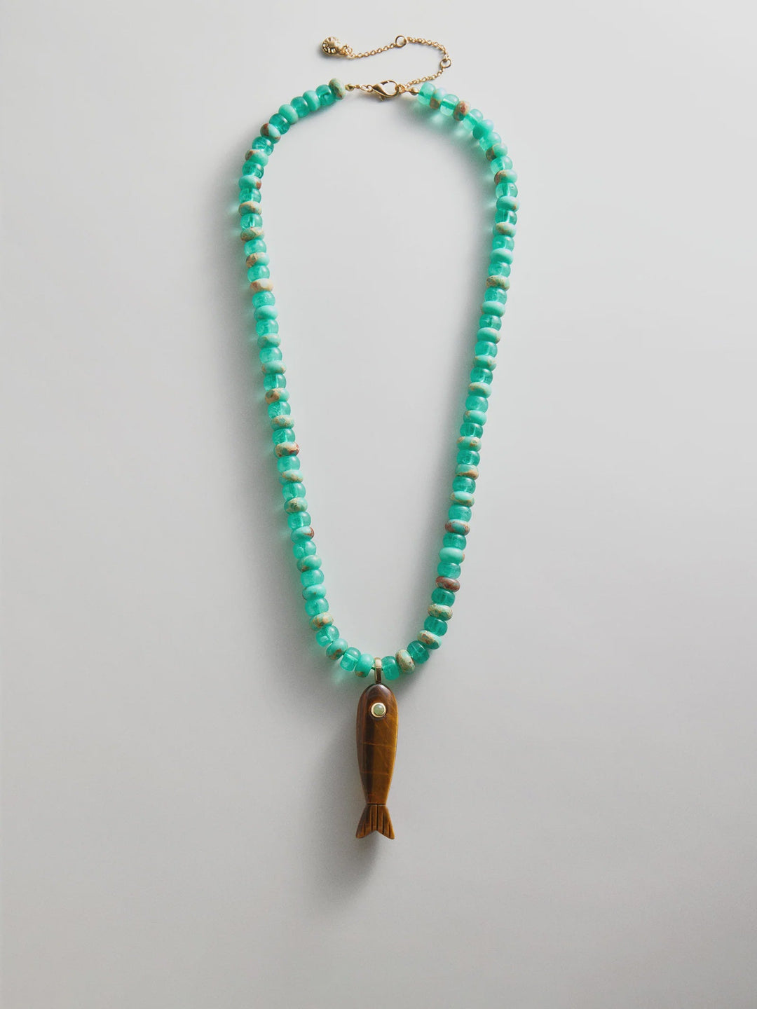 The Zertz SUN DRIFT SEMI-PRECIOUS PNDT NECK features turquoise beads, an adjustable gold clasp, and a brown fish-shaped pendant with a white eye, displayed in an oval shape against a white background.