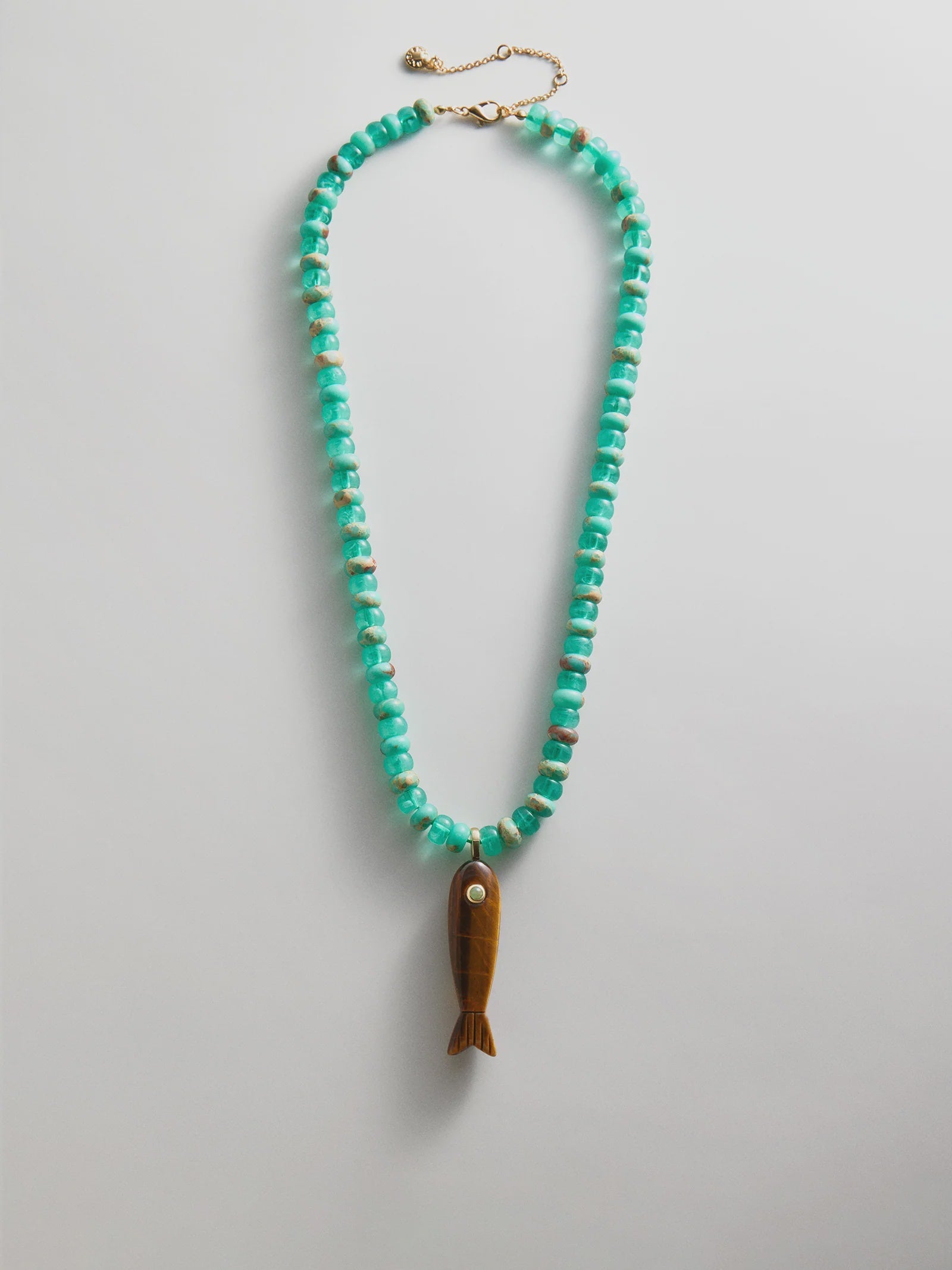 The Zertz SUN DRIFT SEMI-PRECIOUS PNDT NECK features turquoise beads, an adjustable gold clasp, and a brown fish-shaped pendant with a white eye, displayed in an oval shape against a white background.
