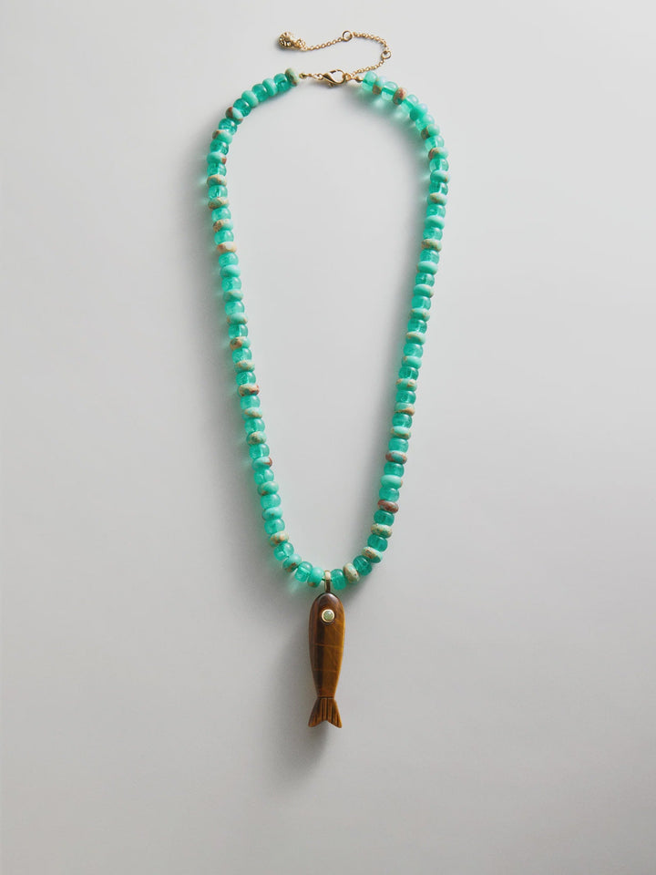 The Zertz SUN DRIFT SEMI-PRECIOUS PNDT NECK features turquoise beads, an adjustable gold clasp, and a brown fish-shaped pendant with a white eye, displayed in an oval shape against a white background.