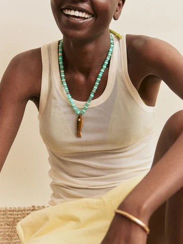 A smiling person in a sleeveless sheer white top wears the Zertz SUN DRIFT SEMI-PRECIOUS PNDT NECK, paired with a light yellow skirt and simple gold bracelet, sitting with one arm on their knee.