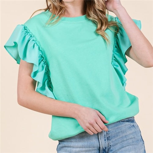 A woman models the Loca on Main Rufflina Top in mint green with short sleeves and ruffled shoulders, paired with light blue jeans, standing against a light beige background. Her face is partially out of frame.