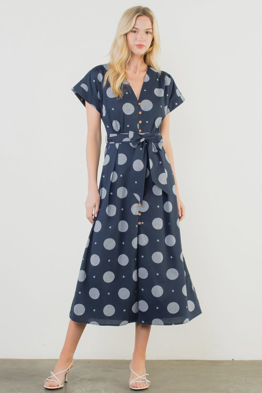 A woman models the Zertz SHORT SLV POLKA DOT PATTERN DRESS—navy, V-neck, button-up, matching waist tie—paired with white strappy sandals against a plain white background.