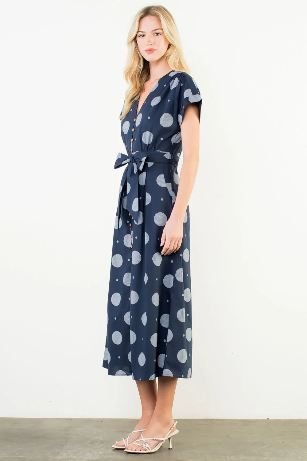 A woman models the Zertz SHORT SLV POLKA DOT PATTERN DRESS—a navy midi with short sleeves, button-down front, and waist tie—paired with white strappy heels, standing against a plain white background.