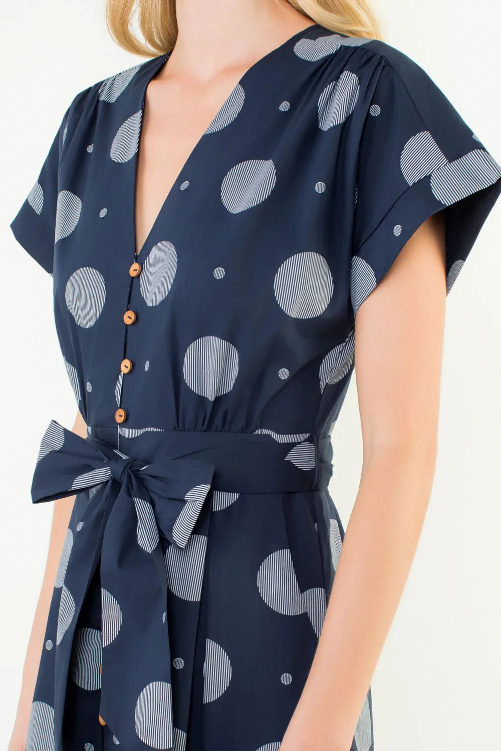 A woman wears the Zertz SHORT SLV POLKA DOT PATTERN DRESS, featuring short sleeves, a deep V-neck, tan buttons, a waist tie, and bold white polka dots—some detailed with fine stripes.