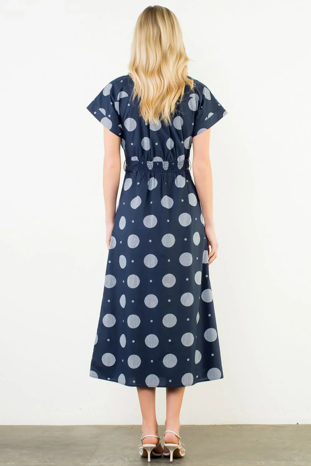 A woman with long blonde hair faces away, wearing the Zertz SHORT SLV POLKA DOT PATTERN DRESS, a navy cotton midi dress with short sleeves, paired with white heeled sandals. The background is plain white.