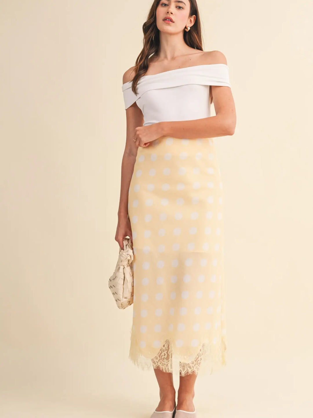 A woman wearing Zertz’s POLKA DOT LACE TRIM SKIRT and a white off-the-shoulder top, holding a small bag, stands against a beige background.