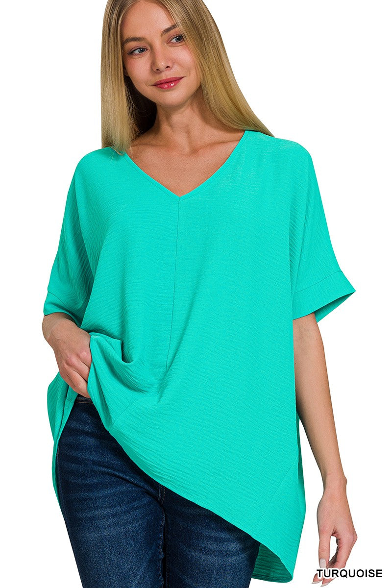 A woman with long blonde hair wears the Loca on Main Airflow Basics Top in turquoise, an oversized, lightweight summer top with a V neckline, short sleeves, and asymmetrical hem. She pairs it confidently with dark blue jeans.