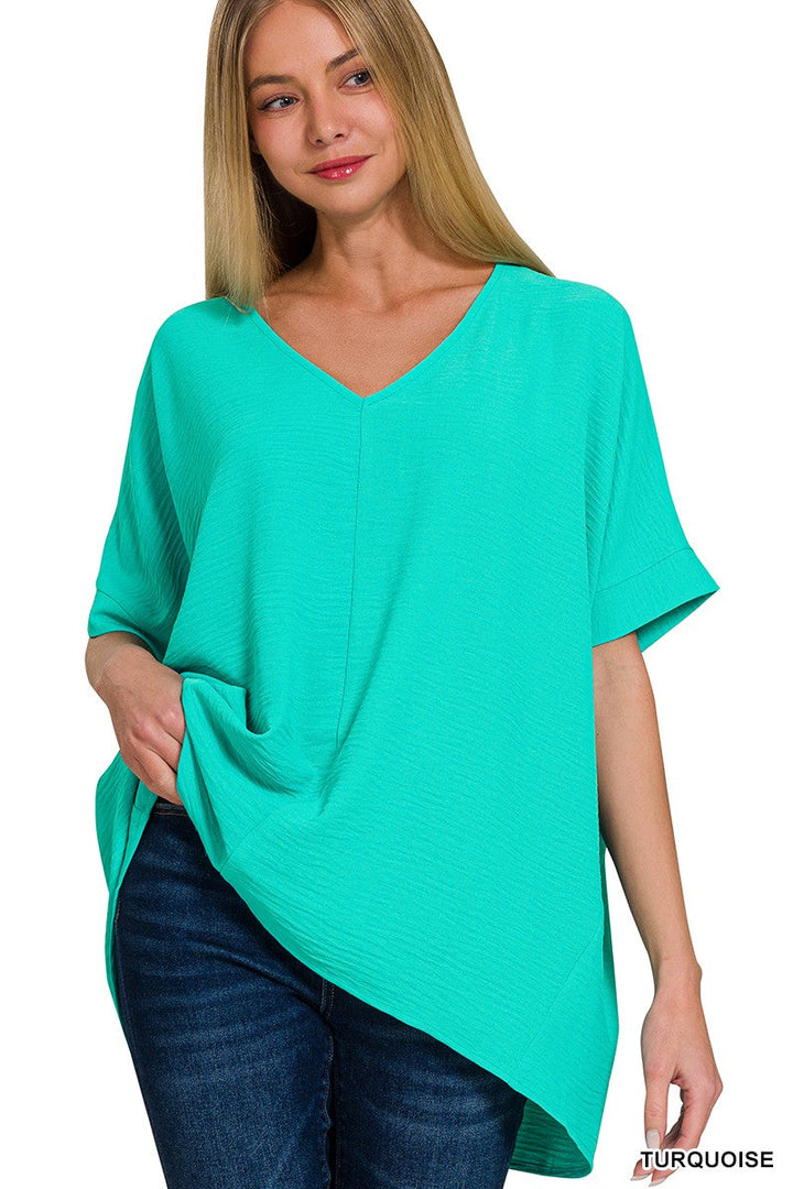 A woman with long blonde hair wears the Loca on Main Airflow Basics Top in turquoise, an oversized, lightweight summer top with a V neckline, short sleeves, and asymmetrical hem. She pairs it confidently with dark blue jeans.