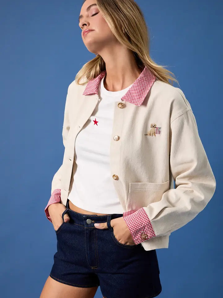 A woman poses against a blue background in the Zertz USA FLAG BARN JACKET, a relaxed-fit canvas jacket with a red checkered collar and cuffs, paired with a white tee with a red star and dark denim shorts. Her eyes are closed, head tilted back.