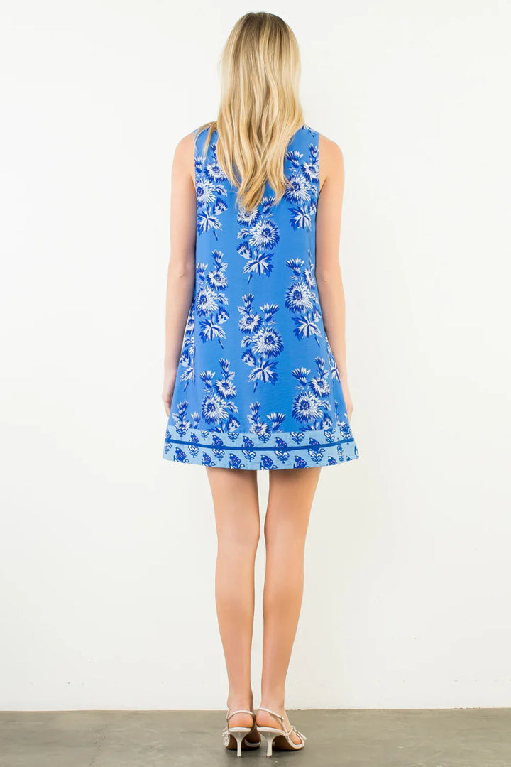 A woman with long blonde hair faces away, wearing the Zertz SLEEVELESS FLORAL PRINT DRESS in blue and white, paired with white heeled sandals. She stands on a concrete floor against a plain white wall.