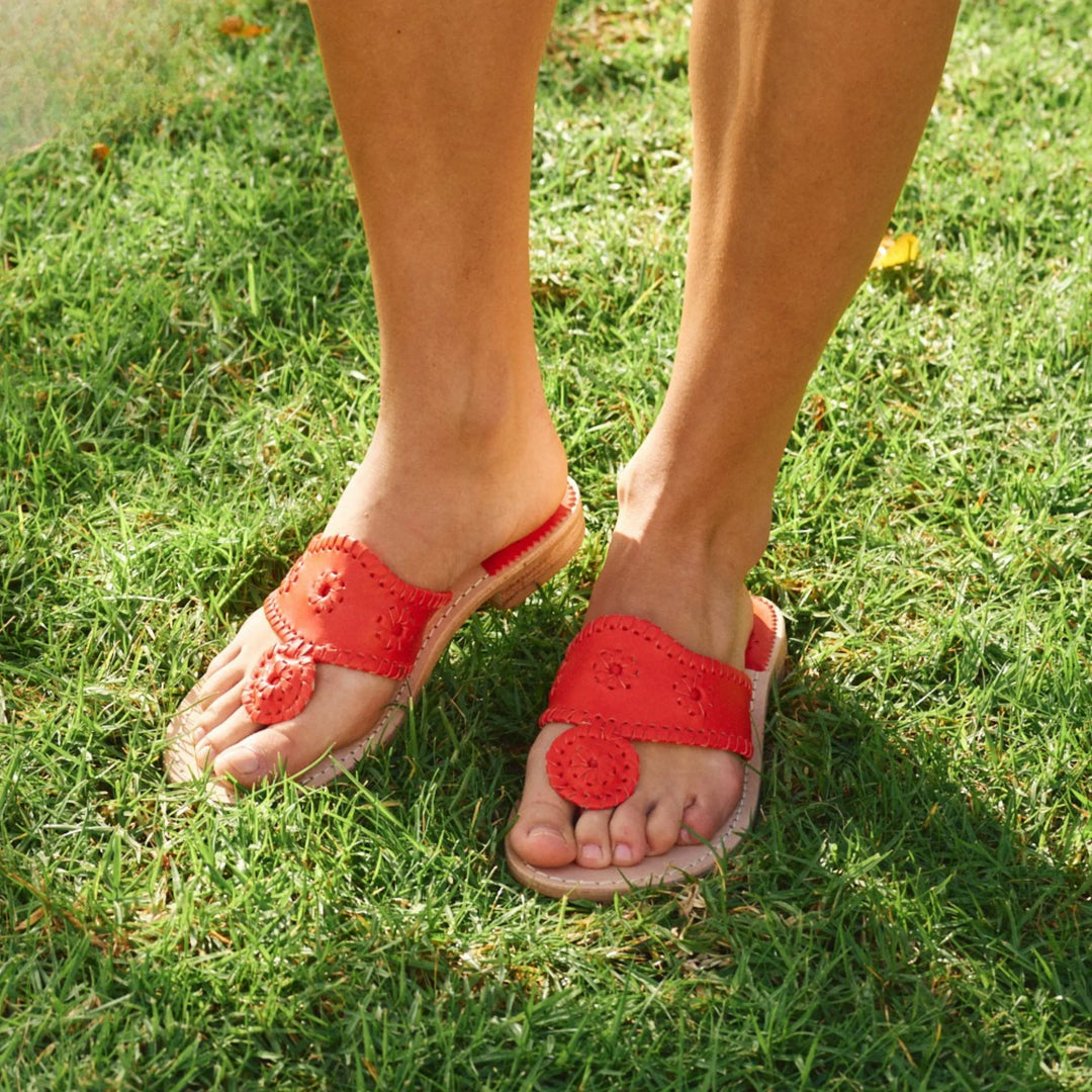 Standing on green grass, a person wears bright red Zertz JACKS FLAT SANDAL NAPPA featuring decorative stitching, cutout strap details, and improved flexibility for all-day comfort.