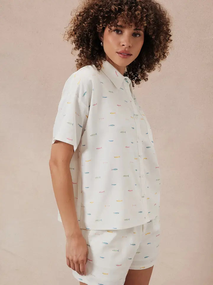 A woman with curly hair models the Zertz PRINTED FISH SHIRT, a short-sleeve button-up, paired with matching white multicolored shorts, evoking ocean-inspired style against a neutral background.