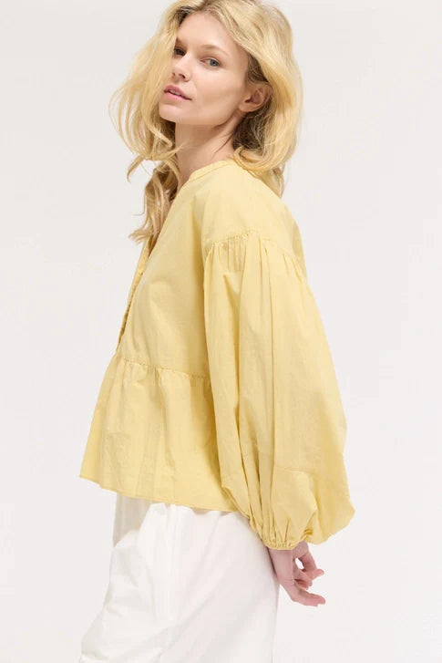 A woman with long blonde hair models the Zertz BALOON SLEEVE BLOUSE in light yellow, paired with white pants, standing sideways against a plain light background—a romantic silhouette for a versatile wardrobe.