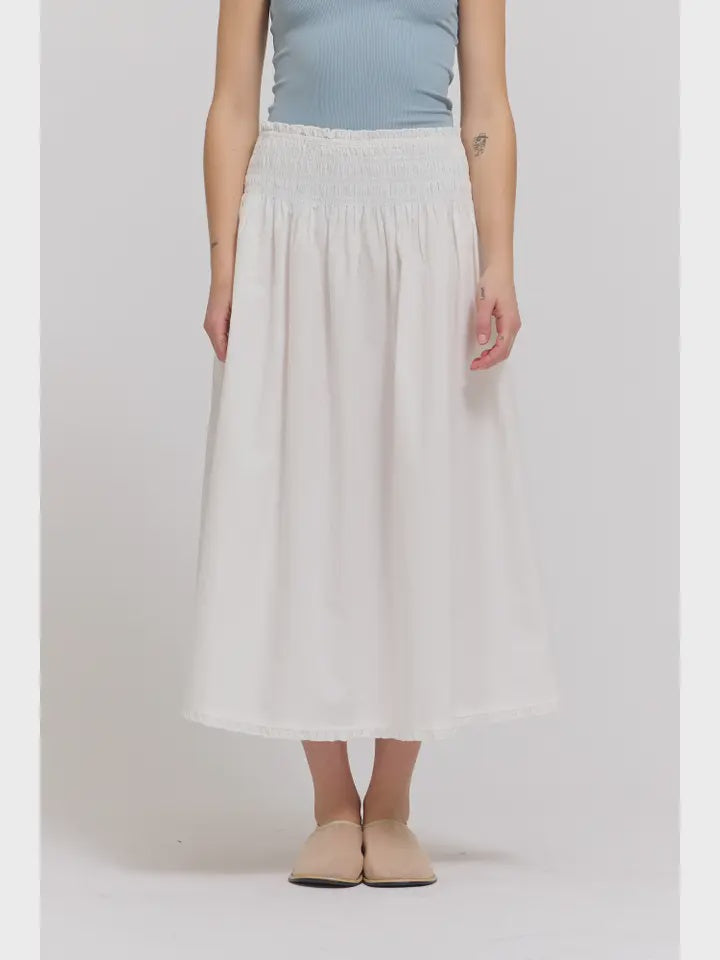 A person wears the Zertz SMOCKED WAIST SKIRT—ankle-length, white, with eyelet trim and detailed design—paired with a light blue sleeveless top and beige flats, standing against a plain light background.